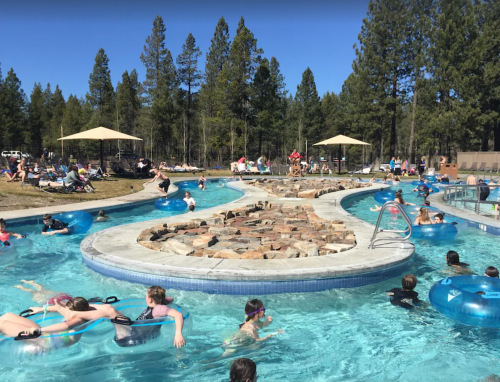 A busy pool area with people floating on inner tubes, surrounded by trees and sun umbrellas on a sunny day.