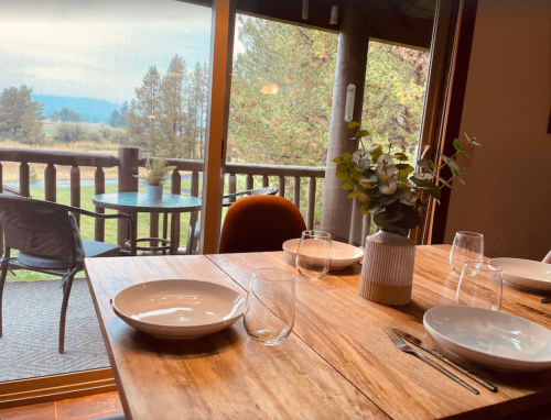 A dining table set with plates and glasses, overlooking a scenic view of trees and mountains through a large window.