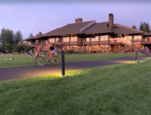 A cyclist rides past a large building at dusk, with people relaxing on the grass nearby.