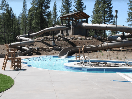 A water park scene featuring a pool, water slides, and lifeguard chairs surrounded by trees and a wooden structure.