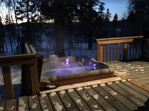A hot tub on a wooden deck surrounded by snow and trees, with purple lights glowing in the evening twilight.
