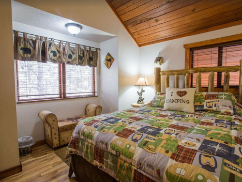 Cozy bedroom with wooden accents, a patterned bedspread, and large windows overlooking a natural setting.