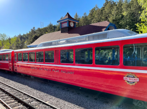A red train at a station with a clock tower and trees in the background, labeled "Pikes Peak Railway."