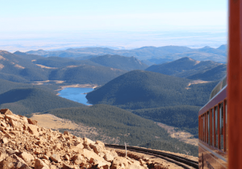 A scenic view from a mountain train, overlooking a lake and rolling hills in the distance under a clear blue sky.