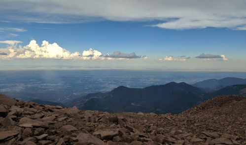A panoramic view from a mountain summit, showcasing rocky terrain and distant valleys under a partly cloudy sky.