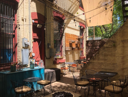 A cozy outdoor café with tables, string lights, and a rustic wall, surrounded by greenery and warm sunlight.