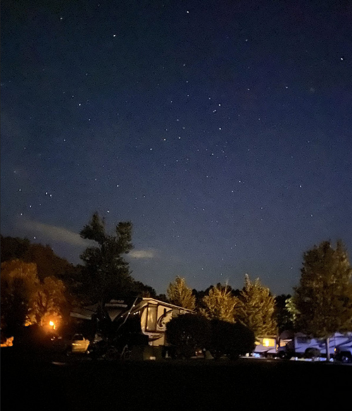 A night sky filled with stars above a campsite, featuring an RV and illuminated trees.