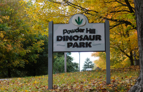 Sign for Powder Hill Dinosaur Park surrounded by colorful autumn foliage.