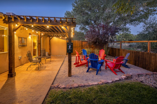 Cozy outdoor seating area with colorful chairs, a fire pit, and string lights, surrounded by greenery and a wooden fence.