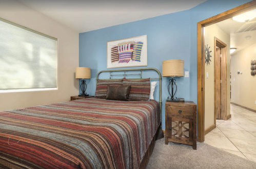 Cozy bedroom with a striped bedspread, two lamps, and a blue accent wall, featuring a hallway in the background.