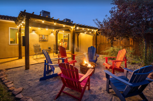 Cozy outdoor space with colorful chairs around a fire pit, string lights, and a patio in the background at dusk.