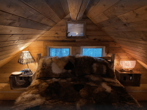 Cozy attic bedroom with a fur-covered bed, wooden walls, and warm lighting from lamps on either side.