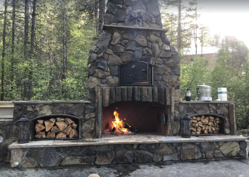 A stone outdoor fireplace with a roaring fire, surrounded by stacked firewood and lush greenery in the background.