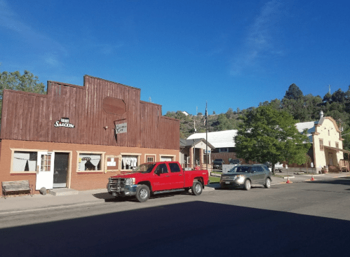 A small town street featuring rustic buildings, a red truck parked, and a backdrop of green hills.