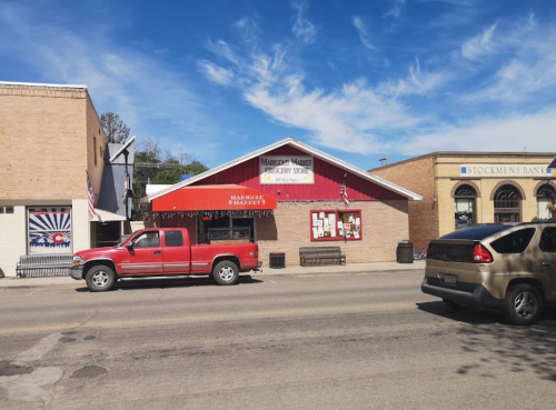 A red-roofed grocery store with a truck parked outside, located on a street with other buildings and a clear blue sky.