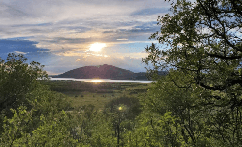 A serene landscape featuring a sunset over a lake, surrounded by lush greenery and distant hills.