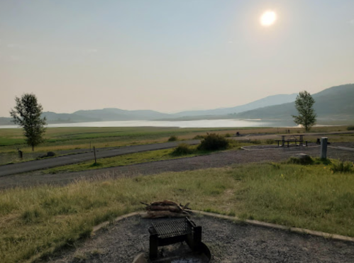 A serene landscape featuring a lake, mountains, and a smoky sky, with a fire pit in the foreground.