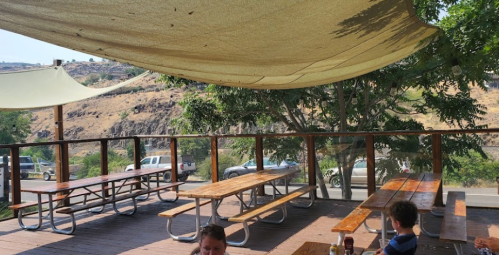 Outdoor dining area with wooden picnic tables shaded by a canopy, surrounded by hills and parked cars.
