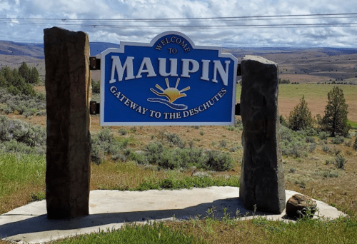 Welcome sign for Maupin, Oregon, featuring the sun and text "Gateway to the Deschutes," surrounded by grassy landscape.