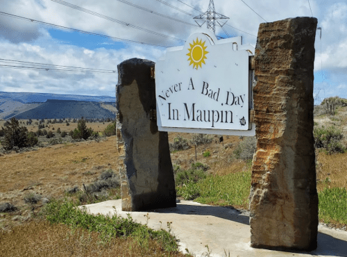 Sign reading "Never A Bad Day In Maupin" surrounded by rocky pillars, with a scenic landscape in the background.
