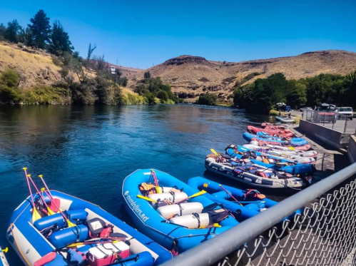 A row of colorful rafts tied up along a riverbank, with hills and trees in the background under a clear blue sky.