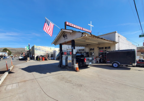A vintage gas station with an American flag, surrounded by vehicles and a clear blue sky.