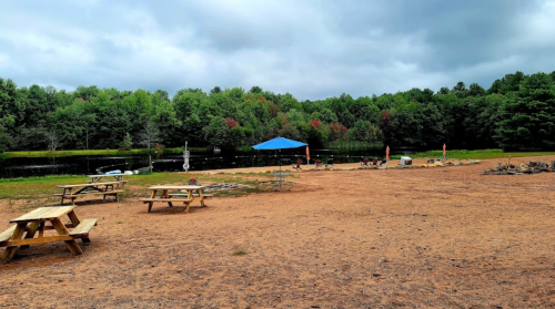 A sandy area by a calm lake, featuring picnic tables and a blue canopy, surrounded by lush green trees.
