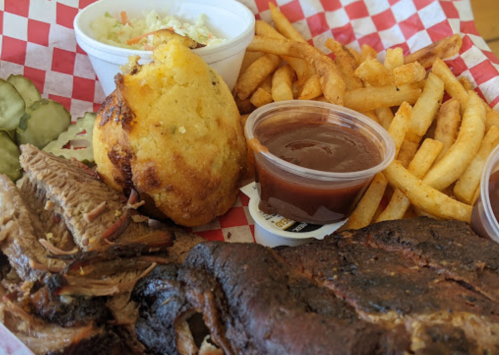 A plate of barbecue with sliced meat, cornbread, fries, coleslaw, pickles, and barbecue sauce in cups, served in a basket.