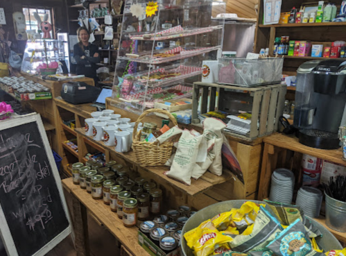 A cozy shop interior filled with jars, snacks, mugs, and baked goods, with a display case and a chalkboard sign.