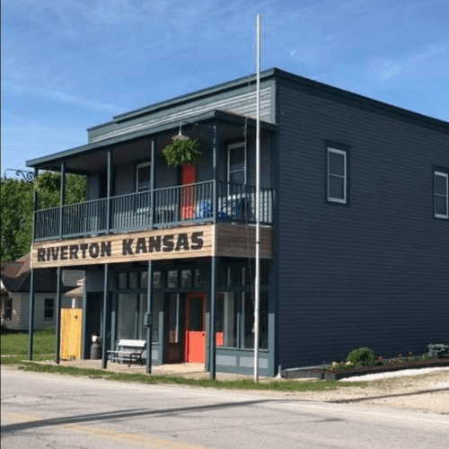 A two-story building with a sign reading "Riverton Kansas," featuring a red door and balcony, set in a small town.