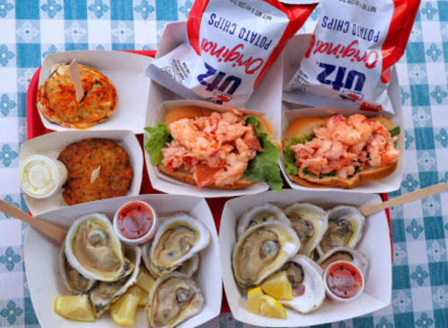 A colorful seafood platter featuring lobster rolls, fried oysters, potato chips, and dipping sauces on a checkered tablecloth.