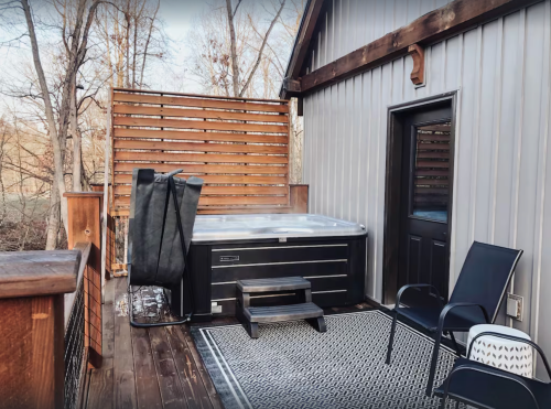 A wooden deck features a hot tub, a black chair, and a privacy screen, surrounded by trees in a serene outdoor setting.