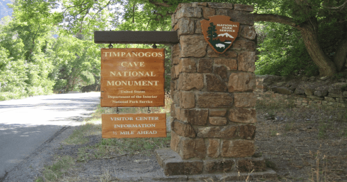 Sign for Timpanogos Cave National Monument, featuring visitor center information and a stone structure surrounded by trees.