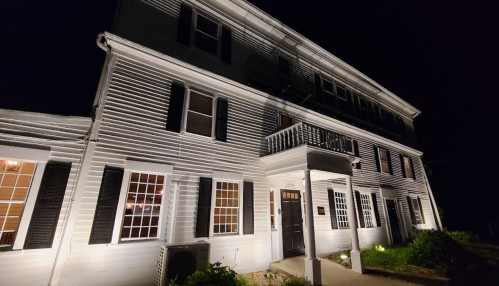 A well-lit, three-story building at night, featuring white siding, multiple windows, and a front porch.