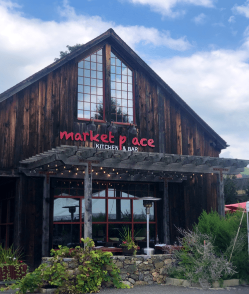 A rustic wooden building with large windows and a sign reading "market place KITCHEN & BAR" under a cloudy sky.