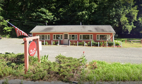 A small, red and gray building with a sign and an American flag, surrounded by greenery and gravel.