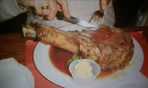 A person cuts into a large, juicy prime rib on a plate, accompanied by a small cup of mashed potatoes.