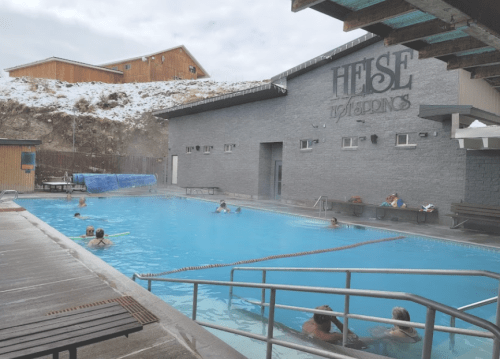 A heated pool with people swimming, surrounded by a building and a hillside under a cloudy sky.