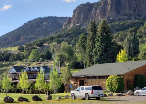 A scenic view of a lodge surrounded by trees and mountains, with a white vehicle parked in the foreground.