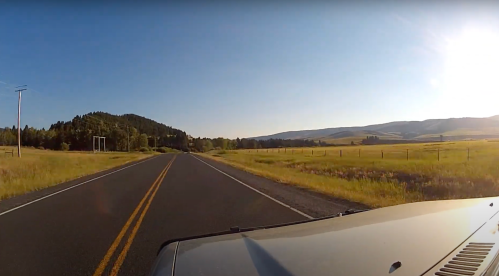 A scenic view of a two-lane road surrounded by fields and mountains under a clear blue sky.