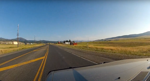A view of a rural road with fields and mountains in the background, captured from a vehicle's perspective.