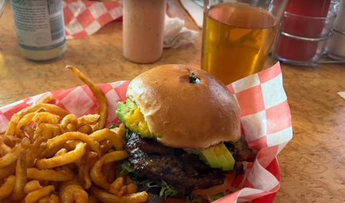 A burger with lettuce, avocado, and a side of curly fries, accompanied by a drink on a table.