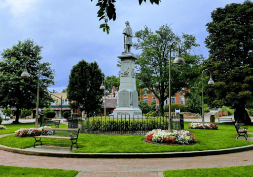 A statue stands in a park surrounded by benches, flowers, and trees under a cloudy sky.