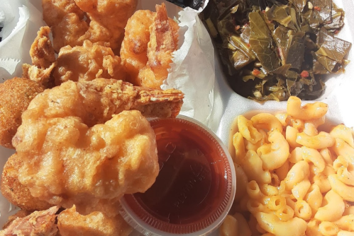 A takeout box with fried shrimp, macaroni and cheese, collard greens, and a small container of sauce.