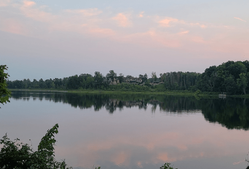 A calm lake reflecting trees and a pastel sky at sunset, with a house visible on the opposite shore.