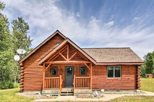 A rustic log cabin with a front porch, surrounded by greenery and under a blue sky with clouds.