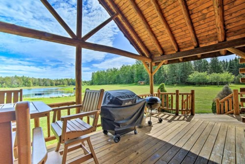 A wooden deck with a grill overlooks a serene lake and lush greenery under a blue sky with scattered clouds.