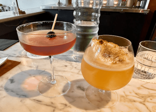 Two cocktails on a marble bar: one in a coupe glass with a cherry, the other in a round glass with a frothy top.