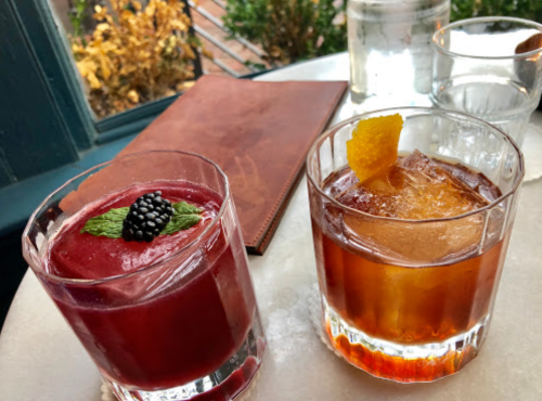 Two cocktails on a table: a berry slush with mint and blackberry, and a whiskey drink with an orange twist.