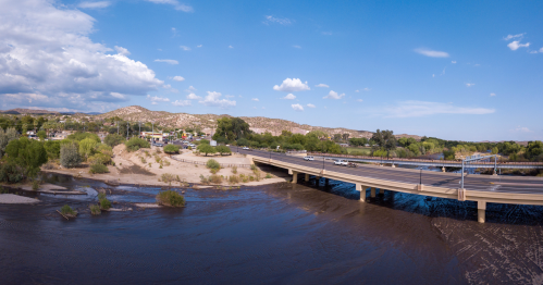 A panoramic view of a river with a bridge, surrounded by greenery and hills under a blue sky with scattered clouds.